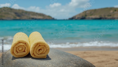Two light yellow towels on a gray rock by a turquoise sea with gentle waves, sandy shore, and blurred rocky hills under a partly cloudy sky—calm, scenic summer beach atmosphere