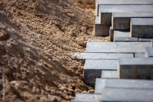 Foto Horizontal close-up perspective showing grey concrete paving stones laid in rows adjacent to a pile of construction sand base during pedestrian walkway installation