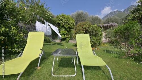 Two green sun loungers in a blooming garden with fresh laundry drying on a line, scenic Italian Alps in the background. Peaceful countryside lifestyle and summer relaxation.