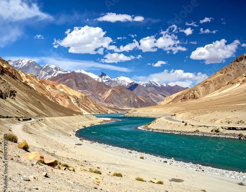 Stunning vista of a vibrant teal river meandering through a valley, flanked by arid landscape and snow-capped mountains under a bright blue sky with fluffy clouds