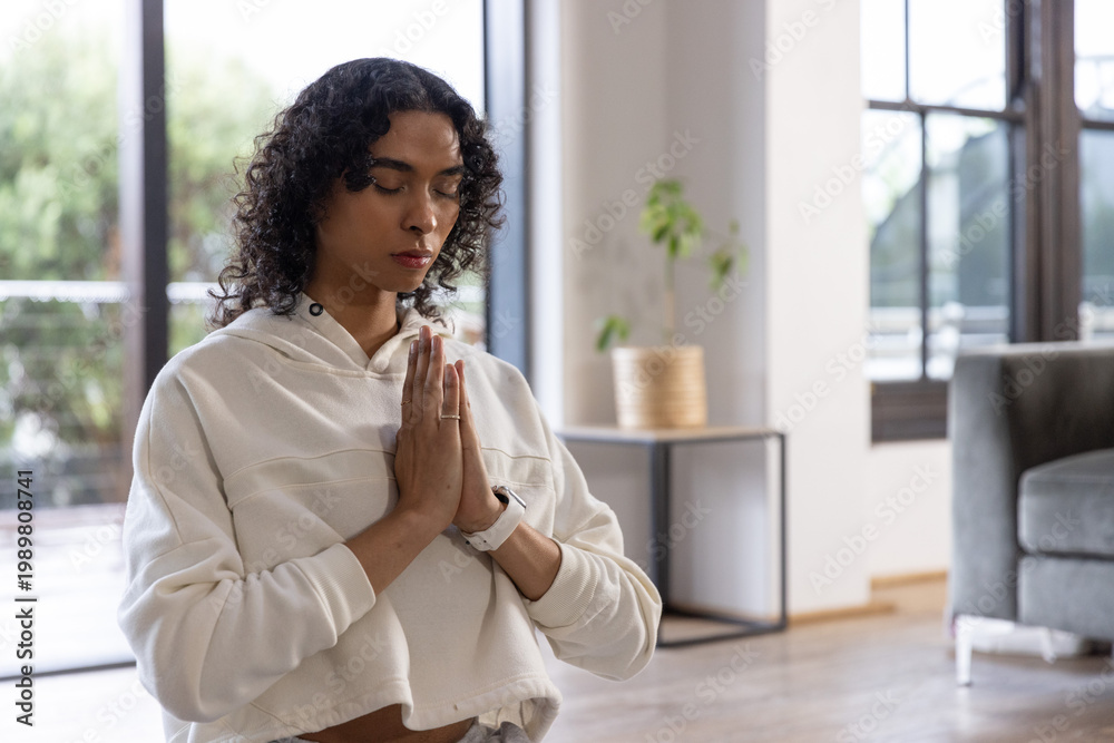 Naklejka premium Non-binary adult meditating with hands in prayer at home on hardwood floor wearing cropped hoodie