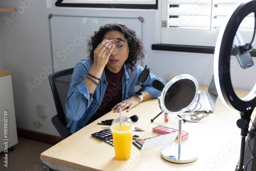 Adult non-binary person applying makeup with cotton pad at light wood desk with mirror, ring light