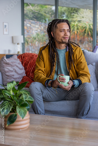 African American man wearing mustard coat sitting on sofa holding green mug looking out window