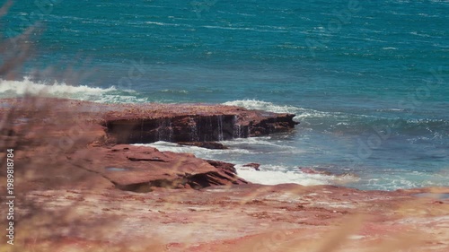 Waves hit the rocks along the shore as water splashes up and sunlight shines lightly across the ocean surface.