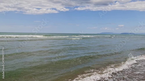 Serene coastal landscape showing small sea waves washing over a sandy shore. Beautiful summer day with blue sky, fluffy clouds, and distant mountain range. Perfect relaxing nature background.