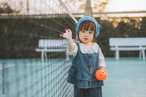 Cute Asian toddler girl holding an orange ball on a pickleball court at sunset. Happy child in denim outfit learning sports, promoting active lifestyle, wellness, and childhood development