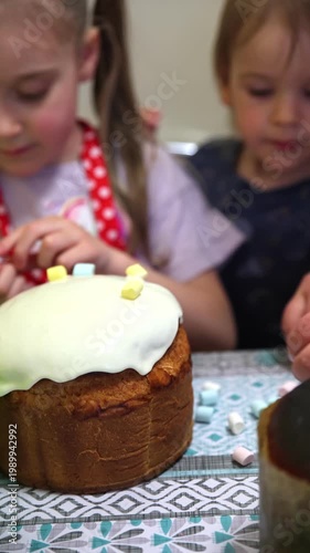Two cute little girls decorating a traditional Easter kulich covered with white icing. The older girl carefully places colorful mini marshmallows on top while the younger one watches with interest.