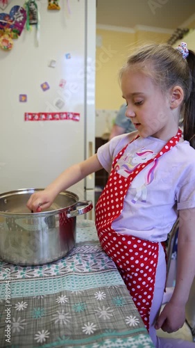 Adorable young girl wearing a red polka-dot apron stands at the table and carefully stirs a big stainless steel pot. Cozy home kitchen scene with refrigerator and colorful magnets in the background. 