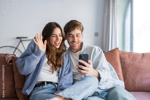 Photos A young couple sits close on a sofa, smiling and waving while video calling on a smartphone, conveying warmth, connection and casual home life in a bright, modern living room