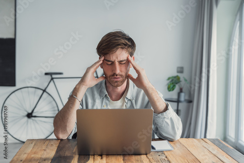 Canvas Print A young man working from home rubs his temples in frustration while staring at a laptop, conveying stress, fatigue and concentration in a modern home office setting with casual decor