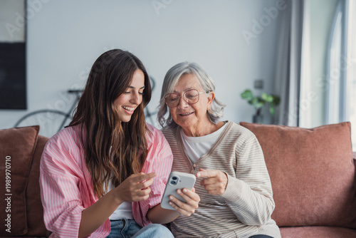 Cuadro en lienzo A young woman helps her elderly grandmother with a smartphone, both smiling and engaged while sitting on a cozy living room couch, sharing a warm family moment and learning technology together