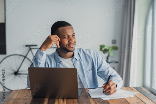 Фотография Thoughtful man working from home, writing notes while using a laptop in a bright, casual home office
