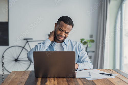 Photos Male remote worker rubs his neck in discomfort while using a laptop at a home office desk with papers, showing fatigue, poor posture and stress during a long work session