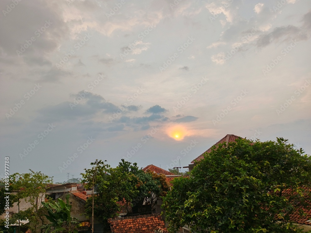 Obraz premium Sunset Over Residential Area with Warm Evening Light, Warm sunset view over houses and trees with soft clouds in the sky, creating a peaceful evening atmosphere. 