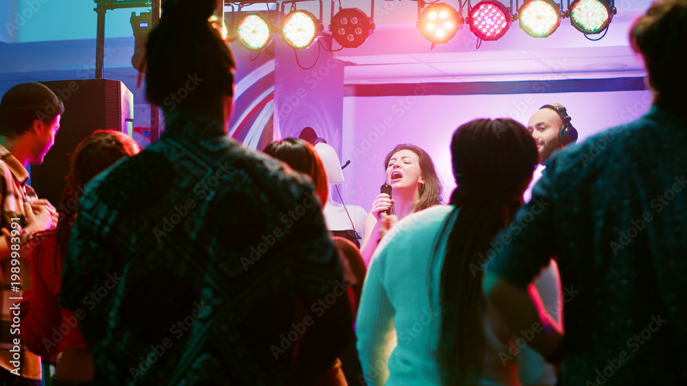 Fototapeta premium Happy woman doing karaoke at party, singing songs with DJ in front of friends on dance floor. Confident girl having fun with live show perfromance next to electronic mixing station at nightclub.