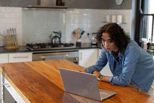 Non-binary adult leaning on wooden island, typing on laptop in kitchen with smartwatch, copy space