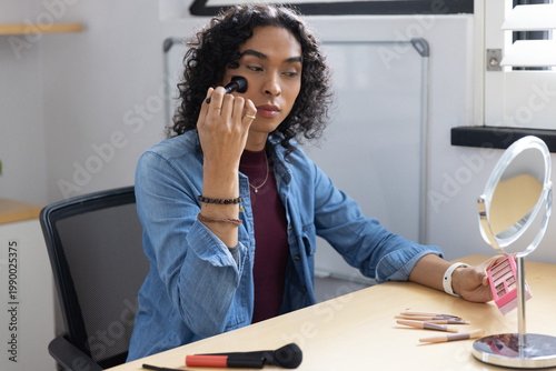 Non-binary adult applying makeup with kabuki brush at wooden desk holding pink palette
