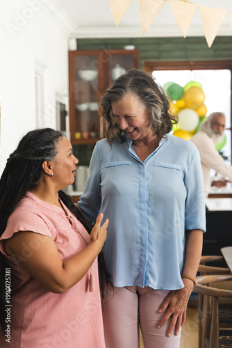 Diverse females in kitchen, left gesturing in pink top, right smiling in blue blouse near balloons