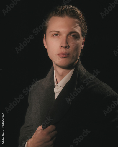 Young blonde man in white dress shirt and charcoal blazer touches collar with direct confident gaze moody dapper refined editorial black studio menswear portrait