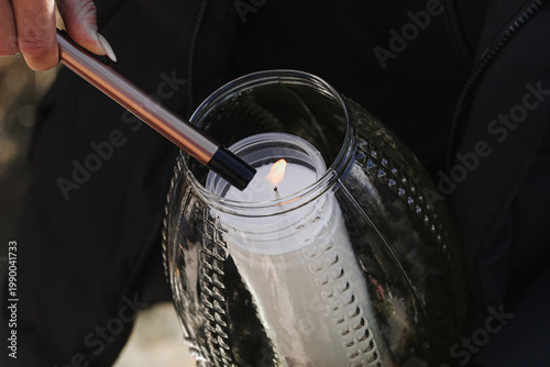 Tablou pe pânză Hand lighting candle inside decorative glass lantern with long lighter outdoors