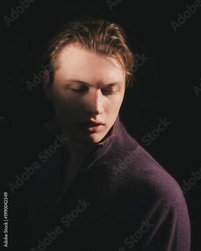 Blonde man in plum purple zip neck sweater lowers head with closed eyes in serene meditative private moment moody delicate studio editorial portrait