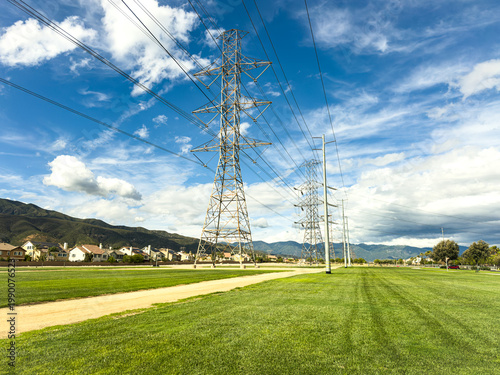 Tall metal power lines stretch across a green field under a bright blue sky with scattered clouds distant mountains and houses line the horizon creating a peaceful landscape