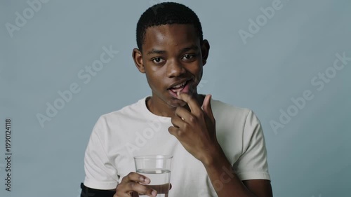 A young African man is holding a glass of cold water; his face betrays his hesitation and fear due to his sensitive tooth enamel