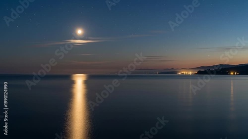 Calm ocean at night under a full moon with distant city lights reflecting on the water.