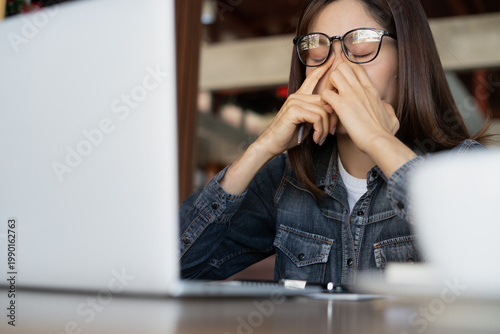 Quadro em tela Stressed woman wearing glasses rubbing eyes and nose while working on laptop in modern indoor workspace