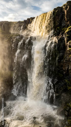 Majestic Waterfall Cascading Over Dark Rocky Cliffs Under Bright Sunlight With Mist Rising From The Turbulent Water Below In A Natural Landscape
