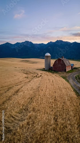 Rustic Red Barn and Silo on a Golden Wheat Field with Majestic Mountains Under a Soft Dusk Sky