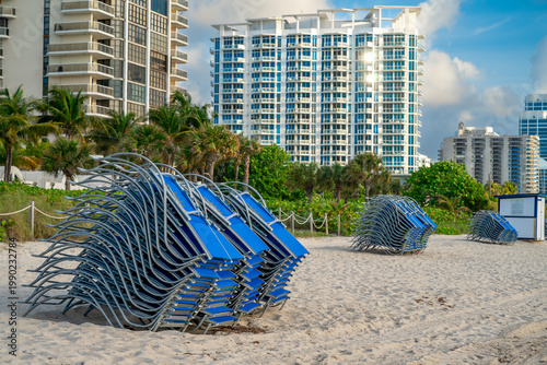 Blue lounge chairs on Miami beach near modern architecture. Close up of stacked sunbeds on sandy beach with white residential buildings and palm trees.