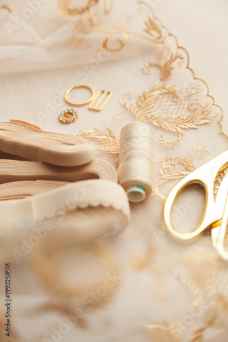 A close-up photo of tailor's tools: beige lace, elastic, spool, accessories for straps, decorations, scissors. Photo grain added.