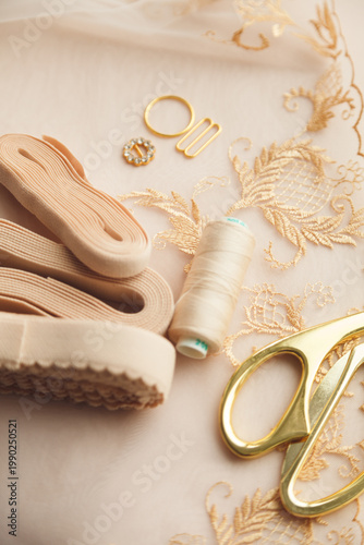 A close-up photo of tailor's tools: beige lace, elastic, spool, accessories for straps, decorations, scissors. Photo grain added.
