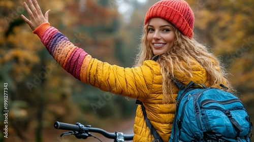 A bright and energetic lifestyle photograph of a smiling young woman with curly blonde hair during an autumn bike ride. She is wearing a yellow puffer jacket, a red beanie, and a backpack