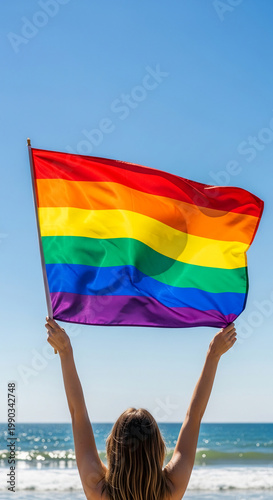 Rear view of a person holding a vibrant rainbow flag high against a clear blue sky and ocean, representing pride and equality