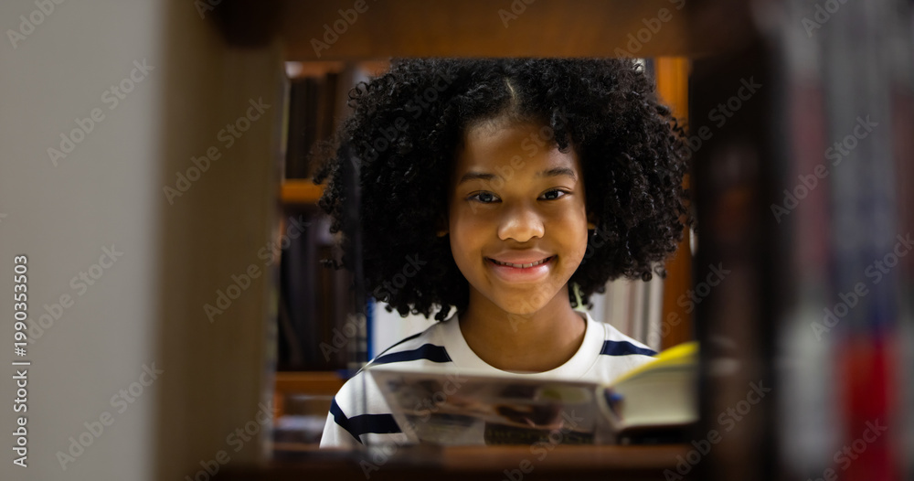 Fototapeta premium Young girl smiles warmly behind bookshelf. Curly hair frames her cheerful expression. She sits in quiet, cozy library space. Image evokes joy, wooden shelves hold many books. Education, Back to school