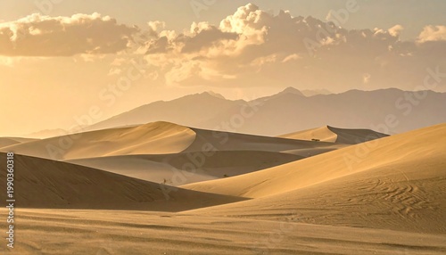 A serene landscape features undulating sand dunes bathed in warm sunlight, with a hazy mountain range in the distance under a cloudy sky