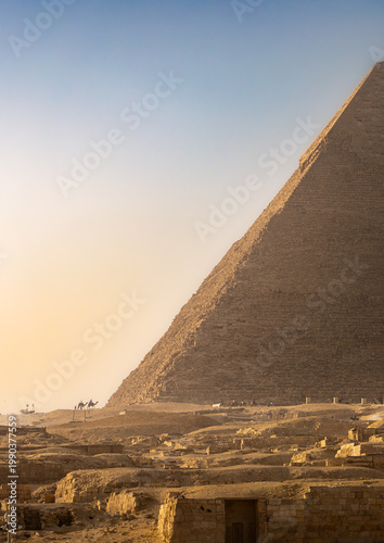 The Great Pyramid of Khufu dominates the frame, its massive limestone blocks rising steeply against a gradient sky transitioning from soft golden haze to clear blue.