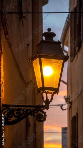 An ornate street lantern glows brightly in a narrow, shadowed alleyway at dusk, against a soft, pastel-colored sky