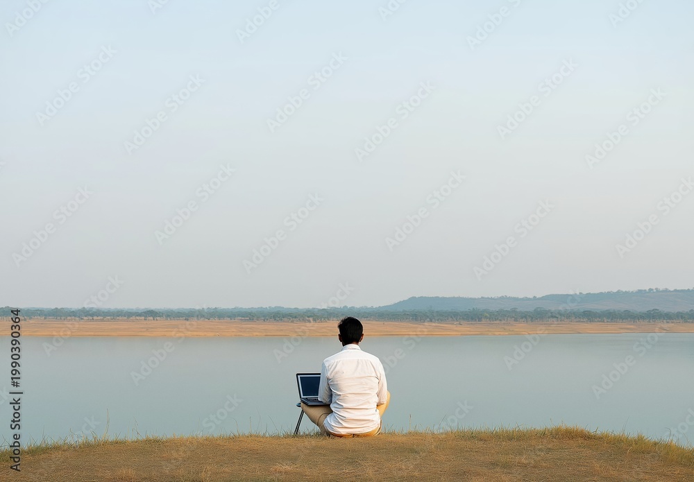 Fototapeta premium Man in casual clothing sitting on a grassy bank, using laptop with a serene lake and hills in the background