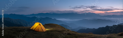 A brightly lit tent sits atop a grassy mountain ridge, overlooking layered blue mountain ranges at twilight. The sky blends with a pink hue