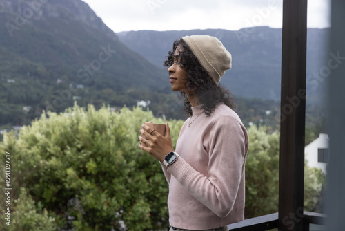 Nonbinary adult standing on balcony holding mug, wearing beanie and smartwatch, gazing at mountains