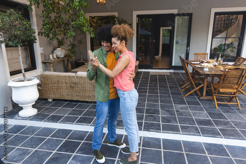 African American couple holding smartphone and smiling on tiled back patio by wooden dining table