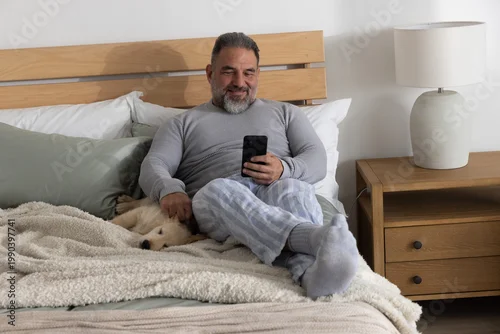 Obraz Mature man sitting on bed with headboard, holding smartphone and petting dog in striped pajamas