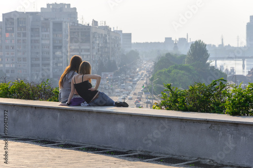 Young girlfriends sitting on a parapet. Cityscape during the day.