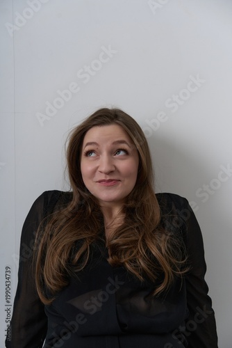 Woman with long hair makes a playful expression while sitting against a white wall in a simple indoor space