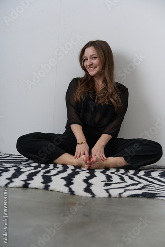 Woman sits on a rug smiling while wearing black clothing indoors in a bright space during daytime