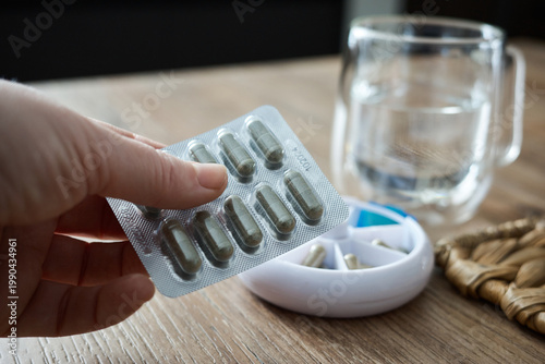 Hand holding a pack of capsules with a glass of water and a pill organizer on a table in a home setting