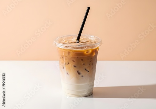 Consider a refreshing iced coffee beverage, complete with a straw, that is lusciously layered with milk and espresso, presented on a table exhibiting a delightfully contrasting peach hue